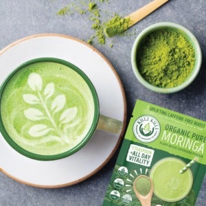 Green moringa latte with a cup of moringa powder and supplements on a table.