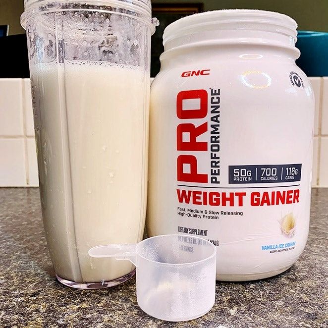 A protein scoop, shaker with a drink and white tub of Pro Weight Gainer.