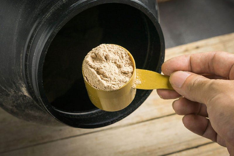 A hand holding a protein scoop with powder, above a black tub.