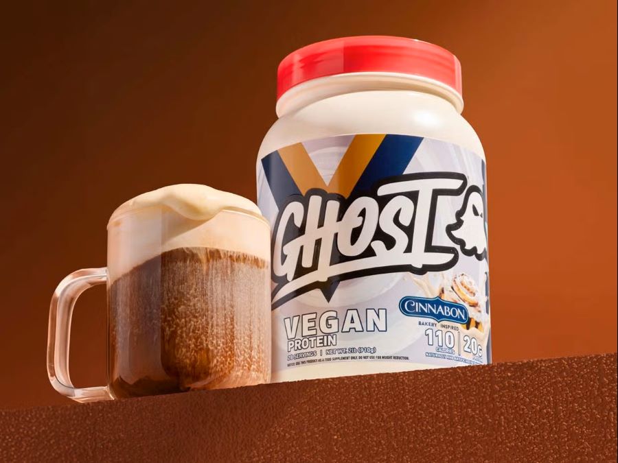 Tub of ghost vegan protein powder next to high protein coffee mug with foam.