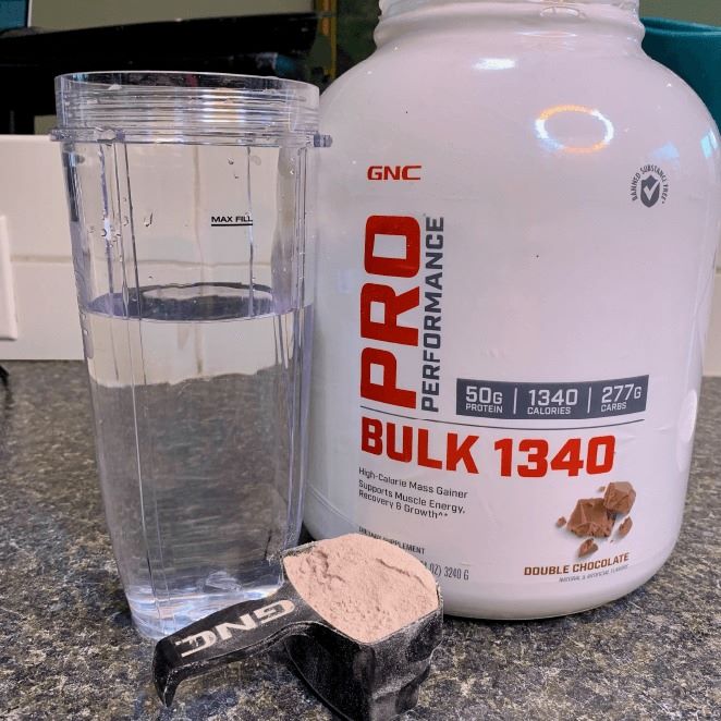 White tub of ProPerformance 1340 Bulk powder, with scoop and cup of water.