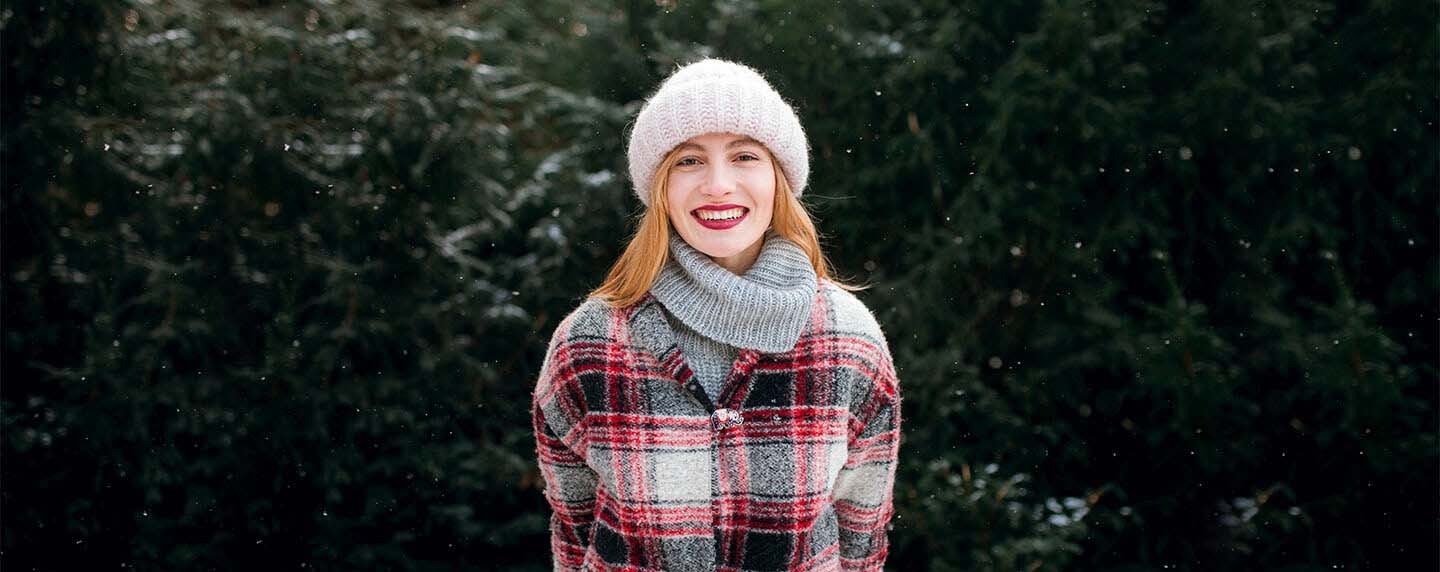 Woman standing outside in snow and smiling wearing hat, scarf and plaid jacket